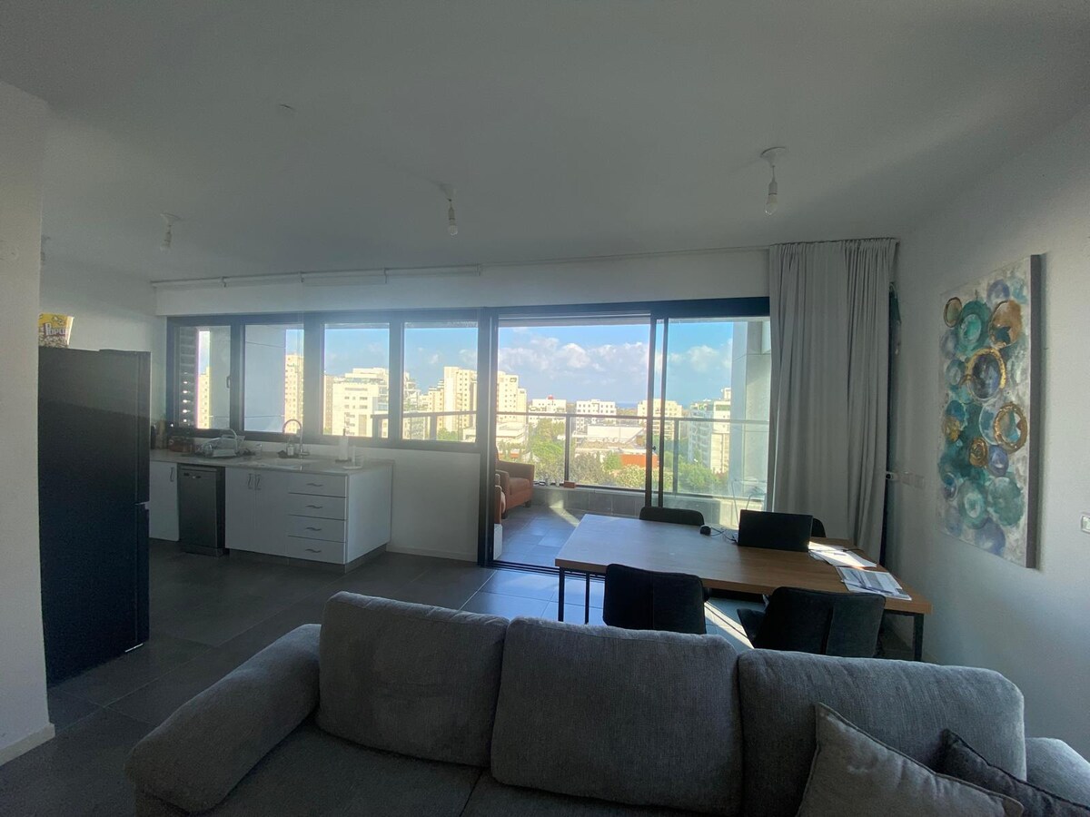 An open living area features large glass doors leading to a balcony with city views. The modern kitchen is equipped with white cabinetry and stainless-steel appliances. A dining table for six is positioned nearby, while a comfortable sofa offers relaxation in the spacious layout.