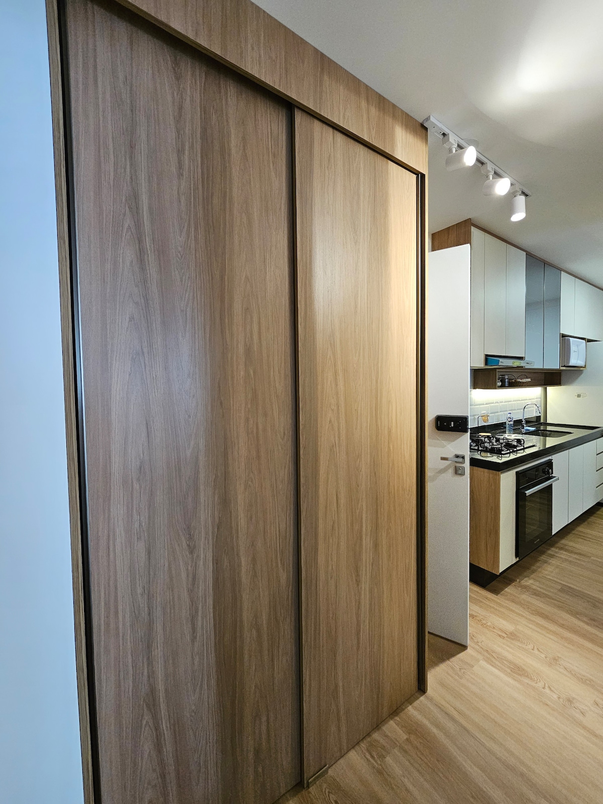 A modern kitchen is displayed, showcasing wooden sliding doors that lead to storage. The interior features sleek cabinetry in light tones, with a gas stove and oven visible, along with countertop space. Ambient lighting enhances the functional atmosphere of the kitchen.
