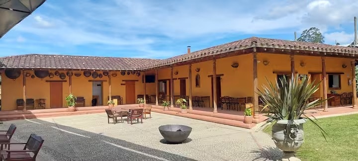 A courtyard is surrounded by a colonial-style building with a terracotta roof. Outdoor seating areas are arranged around the space, complemented by potted plants. A central stone feature adds texture to the scene, while blue skies and gentle clouds create a serene backdrop.