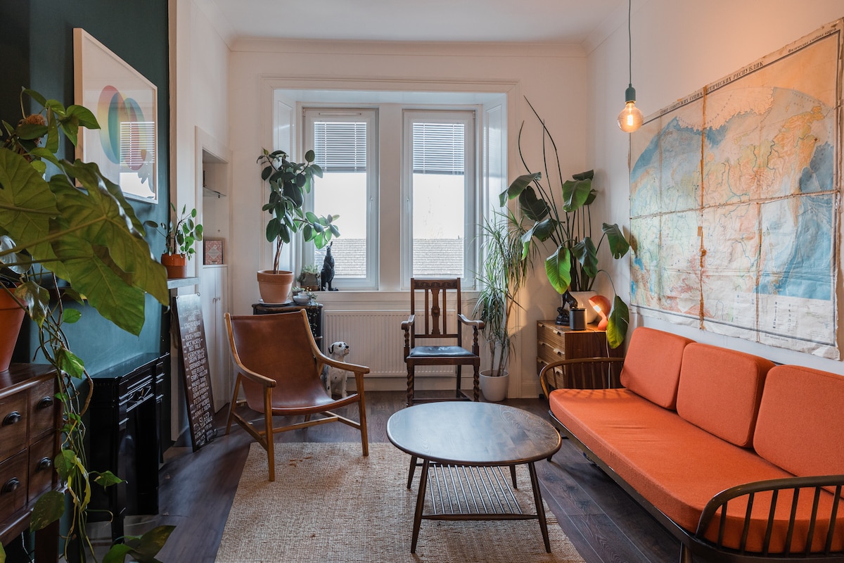 A tastefully arranged living room features a vibrant orange sofa and a round coffee table at its center. Floor plants add greenery, while wall-mounted art and large windows provide natural light, creating a cozy and inviting environment. A wooden chair complements the seating arrangement.