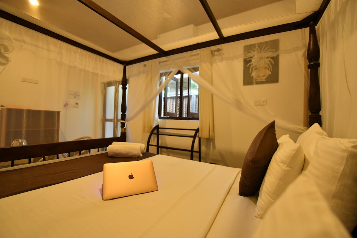A four-poster bed is centered in a bright room, draped with soft white netting. A laptop is placed on the neatly arranged bedding. Light enters through a window, enhancing the space, while decorative wall art adds a touch of character.