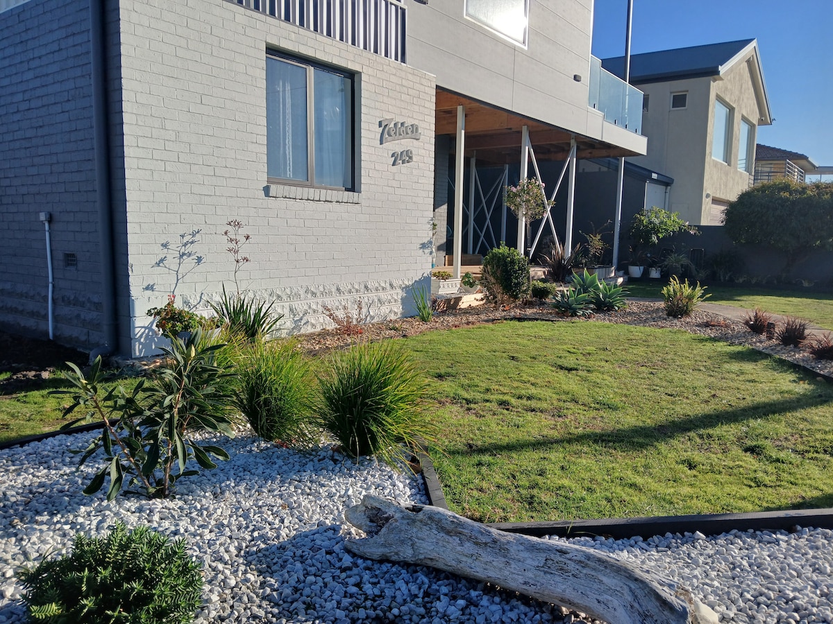 A landscaped front yard features a variety of low-maintenance plants and ornamental grasses, bordered by white gravel. A wooden path leads to the entrance of the separated residence, with a large window reflecting natural light. The overall area is well-kept and inviting.