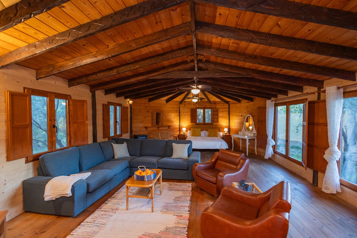A spacious living area in the wooden cabin features a high ceiling with exposed beams and large windows allowing natural light. A comfortable sectional sofa and two armchairs create a welcoming seating arrangement, complemented by a wooden coffee table. A cozy sleeping area is visible in the background.