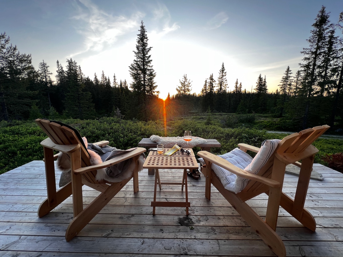 Two wooden chairs are positioned on a deck facing a sunset, surrounded by trees and greenery. A small table sits between them, holding drinks and a book, creating a serene outdoor setting.