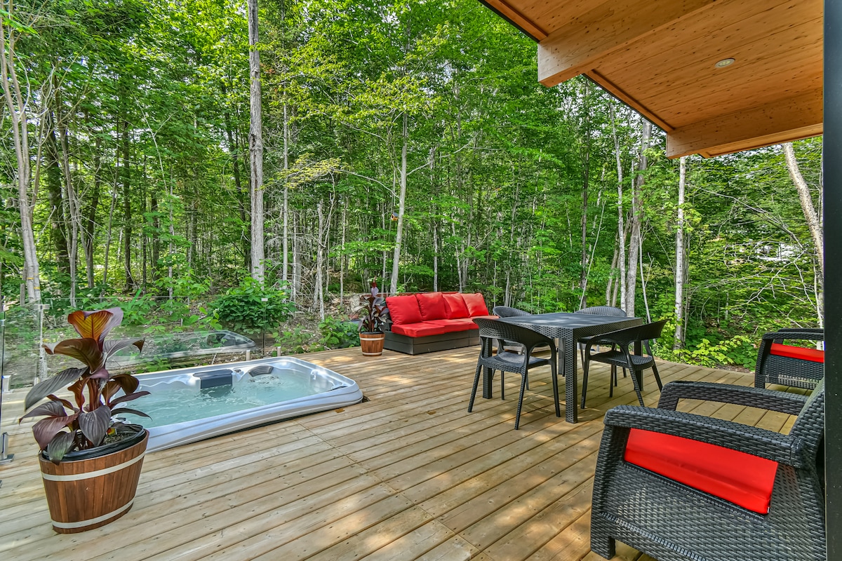 A spacious wooden terrace is surrounded by lush greenery, featuring a hot tub surrounded by planters. A seating area includes a red couch and a dining table. Two black chairs with red cushions are positioned near the edge, enhancing the outdoor relaxation space.