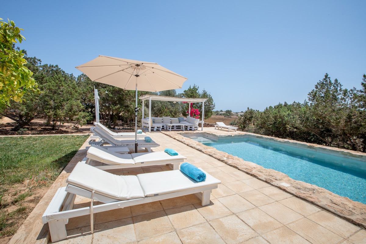 An inviting outdoor space is featured, showcasing a swimming pool surrounded by sun loungers and an umbrella. A shaded seating area with a trellis is visible in the background, set amidst greenery that enhances the tranquil environment.