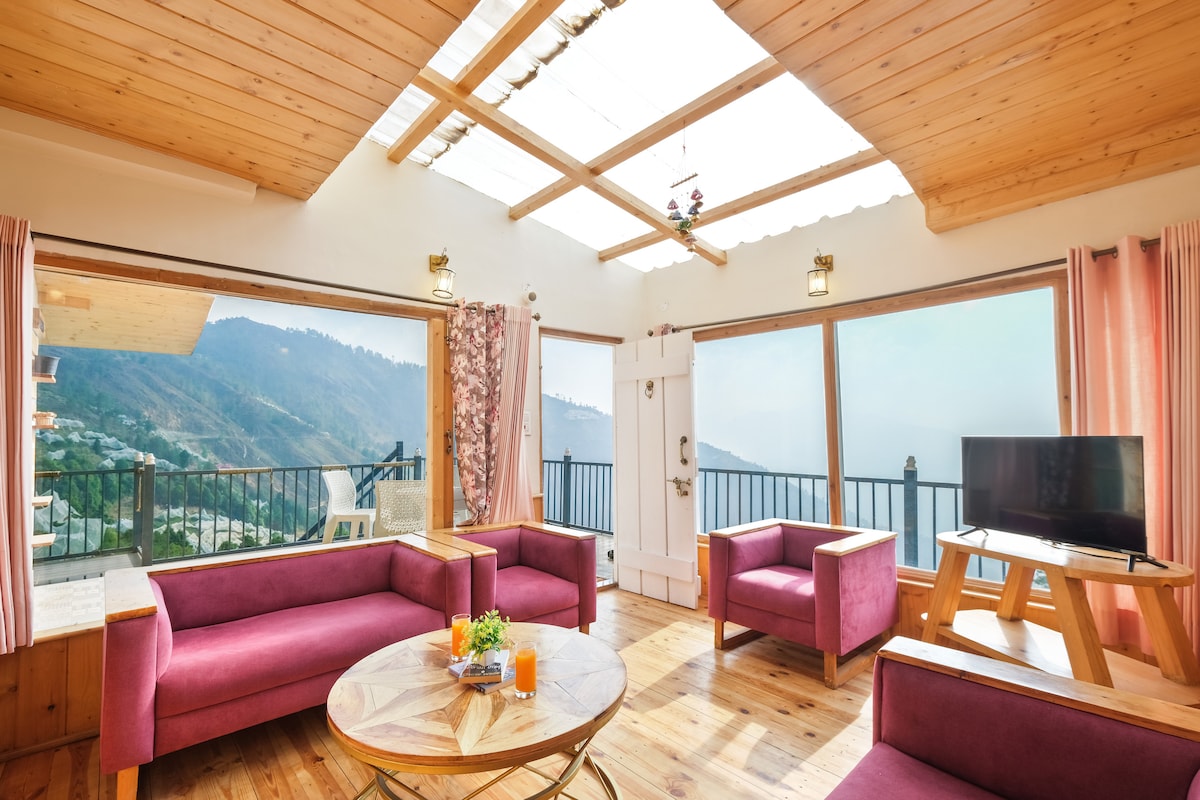 A bright living room is showcased with large windows offering views of the surrounding mountains. Pink armchairs are positioned around a circular wooden coffee table, with a television set visible. Natural light floods the space, highlighting the wooden ceiling and flooring.