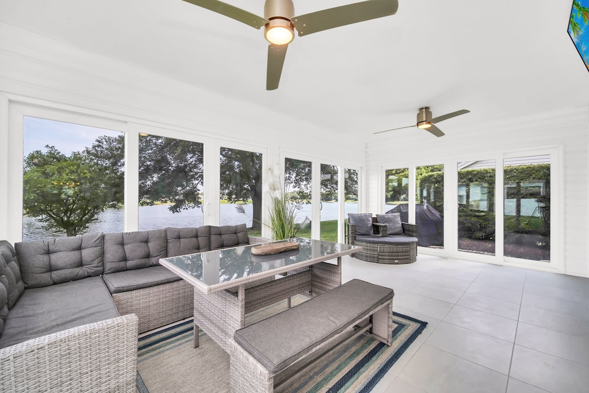 An enclosed lanai showcases comfortable seating arranged around a central glass table, providing views of the waterfront. Large glass doors enhance natural light, while ceiling fans ensure a cool atmosphere. A subtle rug anchors the seating area against the tiled floor.