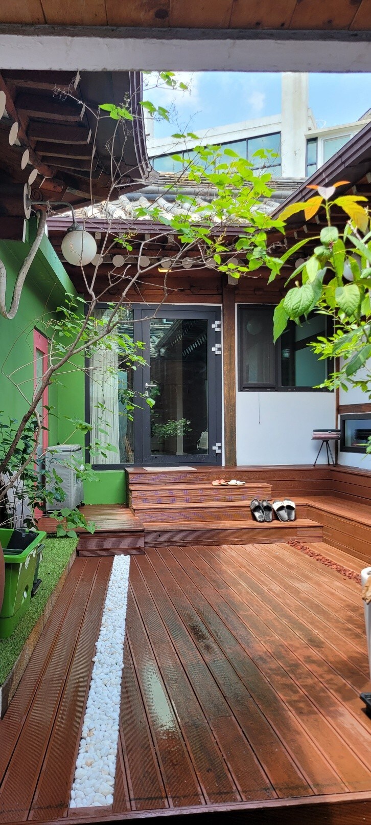 A serene outdoor deck area is provided, featuring wooden flooring and bordered by vibrant greenery. A small stone path leads to a glass door, offering a glimpse of the interior. Two pairs of shoes are placed neatly on the deck, hinting at a welcoming atmosphere.