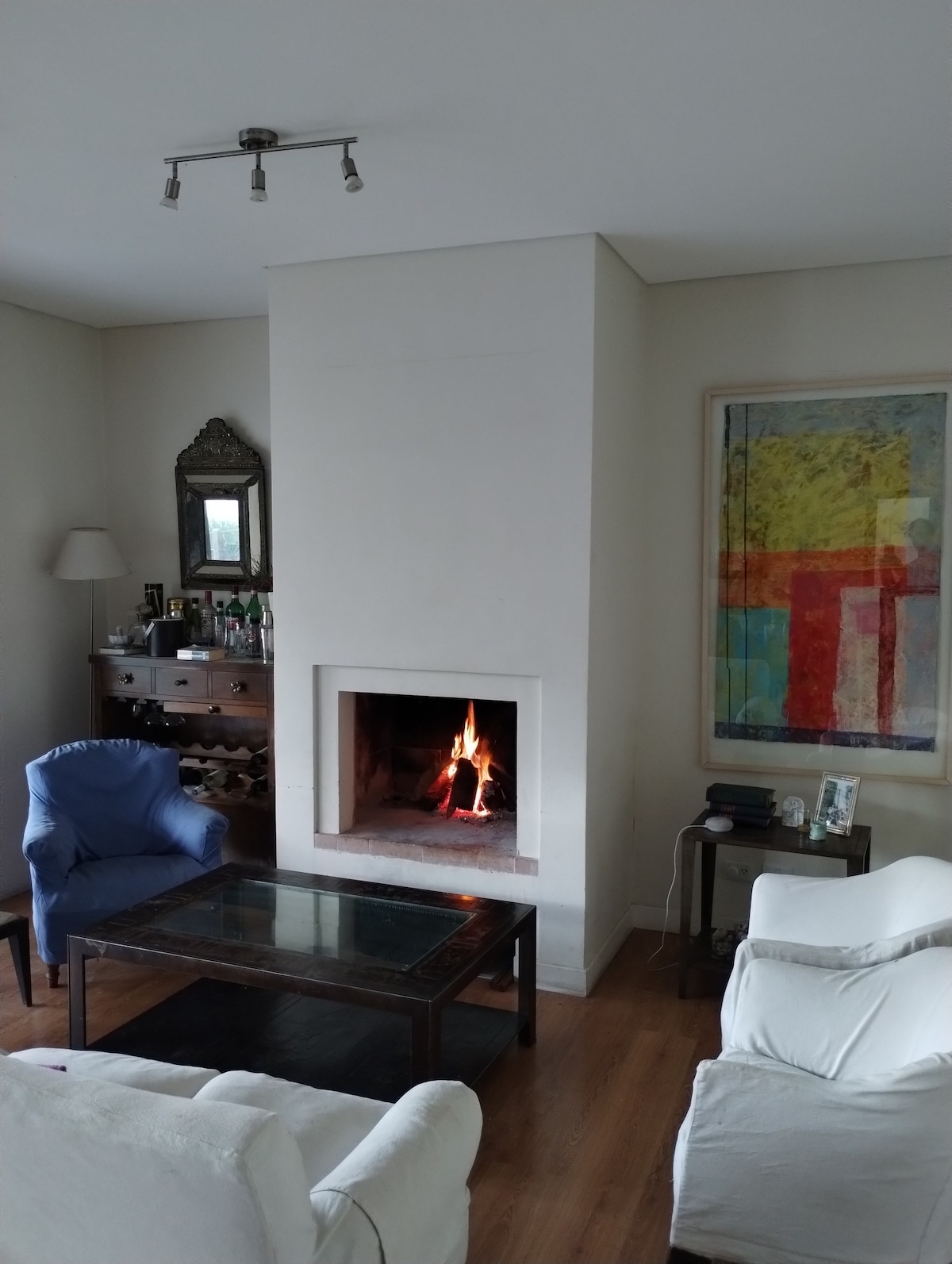 A cozy living area features a modern fireplace with a warm fire, set within a white wall. A glass coffee table rests in the center, surrounded by comfortable seating, including a blue armchair and a white sofa. Artwork adds color to the space, enhancing the overall design.