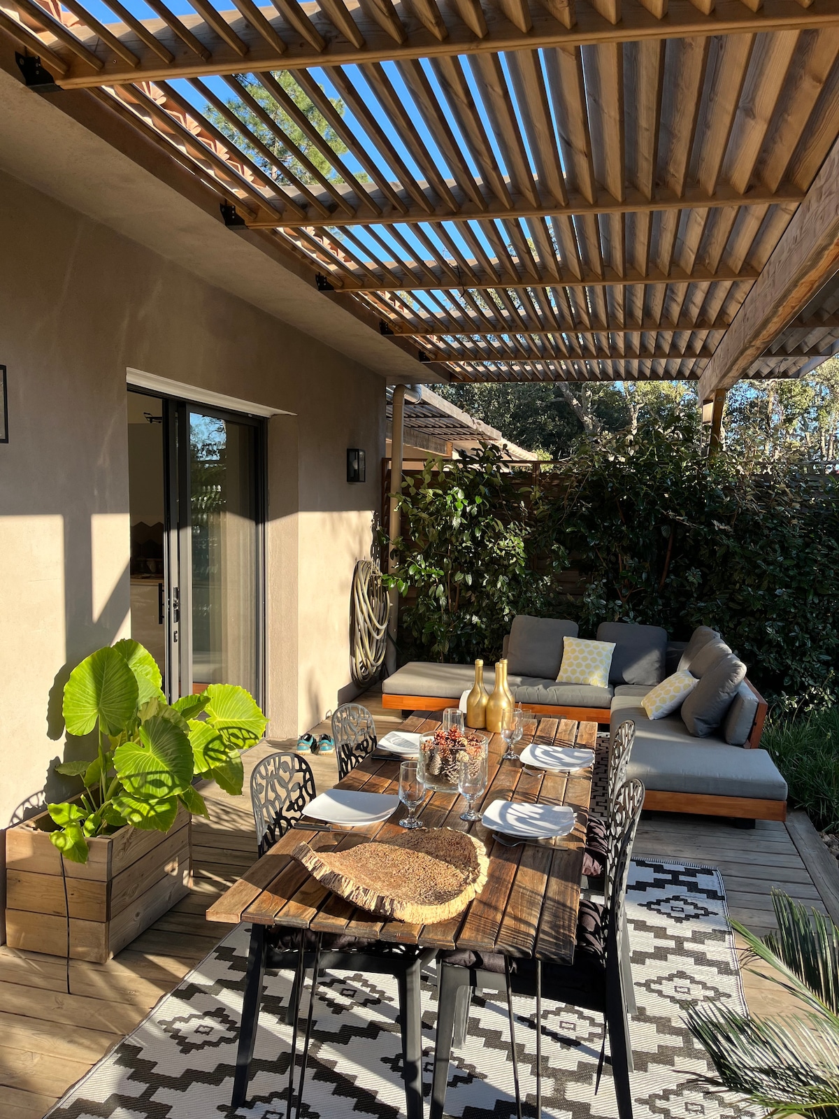 An outdoor terrace is presented with a wooden dining table set for four, featuring a decorative table setting. A comfortable seating area with cushions is located nearby under a wooden pergola, while greenery encircles the space, creating a welcoming atmosphere.