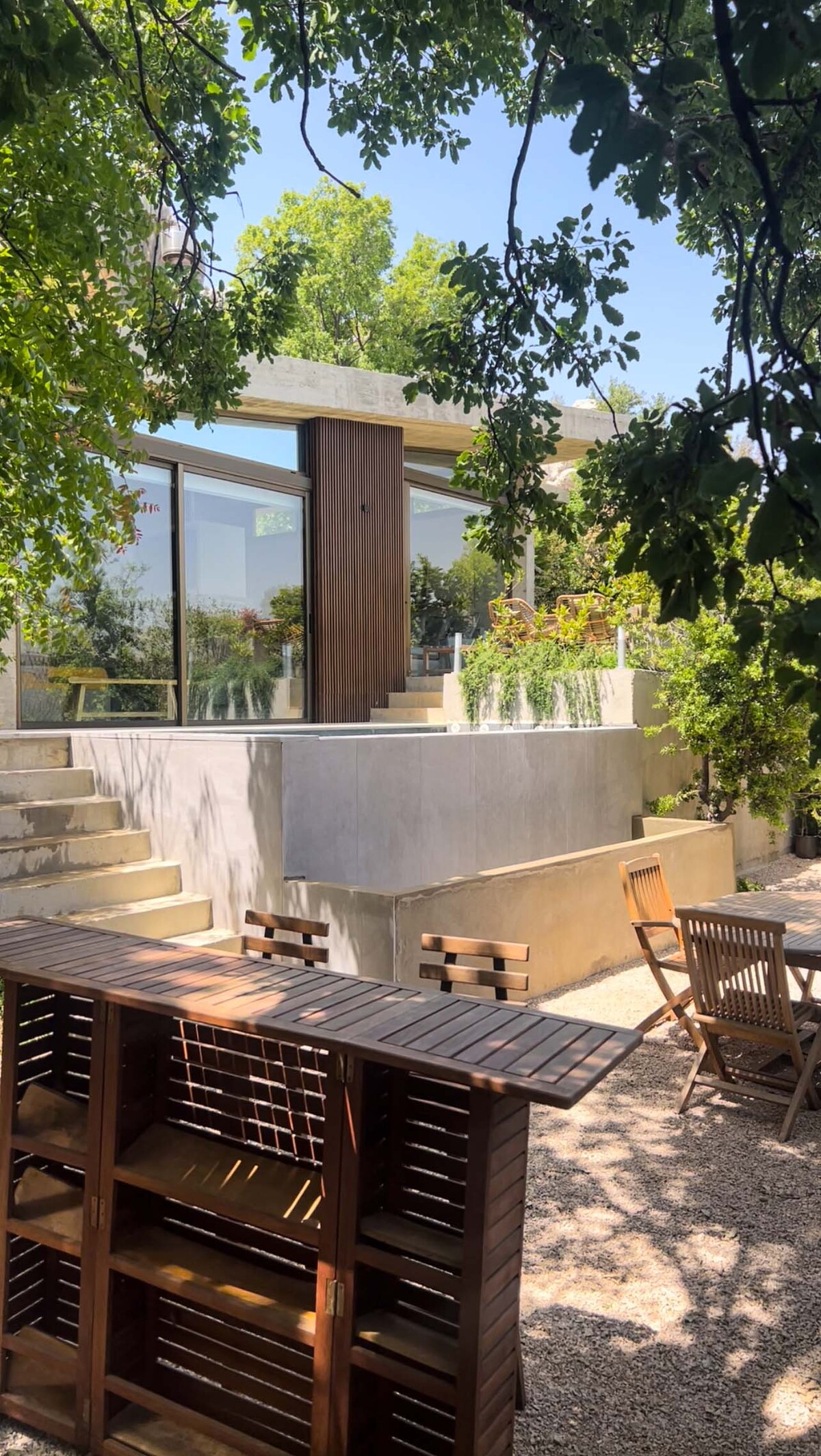 The outdoor area features a wooden table and chairs, surrounded by gravel and greenery. The villa's two-story design is highlighted by large glass panels, inviting natural light. Steps lead to a spacious outdoor terrace where the surroundings are framed by lush foliage.