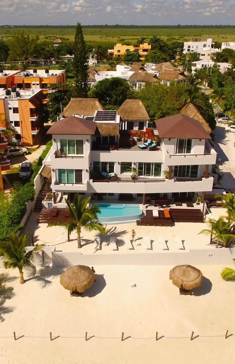 The villa is displayed from an aerial view, showcasing a modern, multi-level structure with a spacious pool area and a private beach. Several lounge chairs are arranged near the pool, and shaded seating areas are visible under thatched cabanas along the sandy shore.