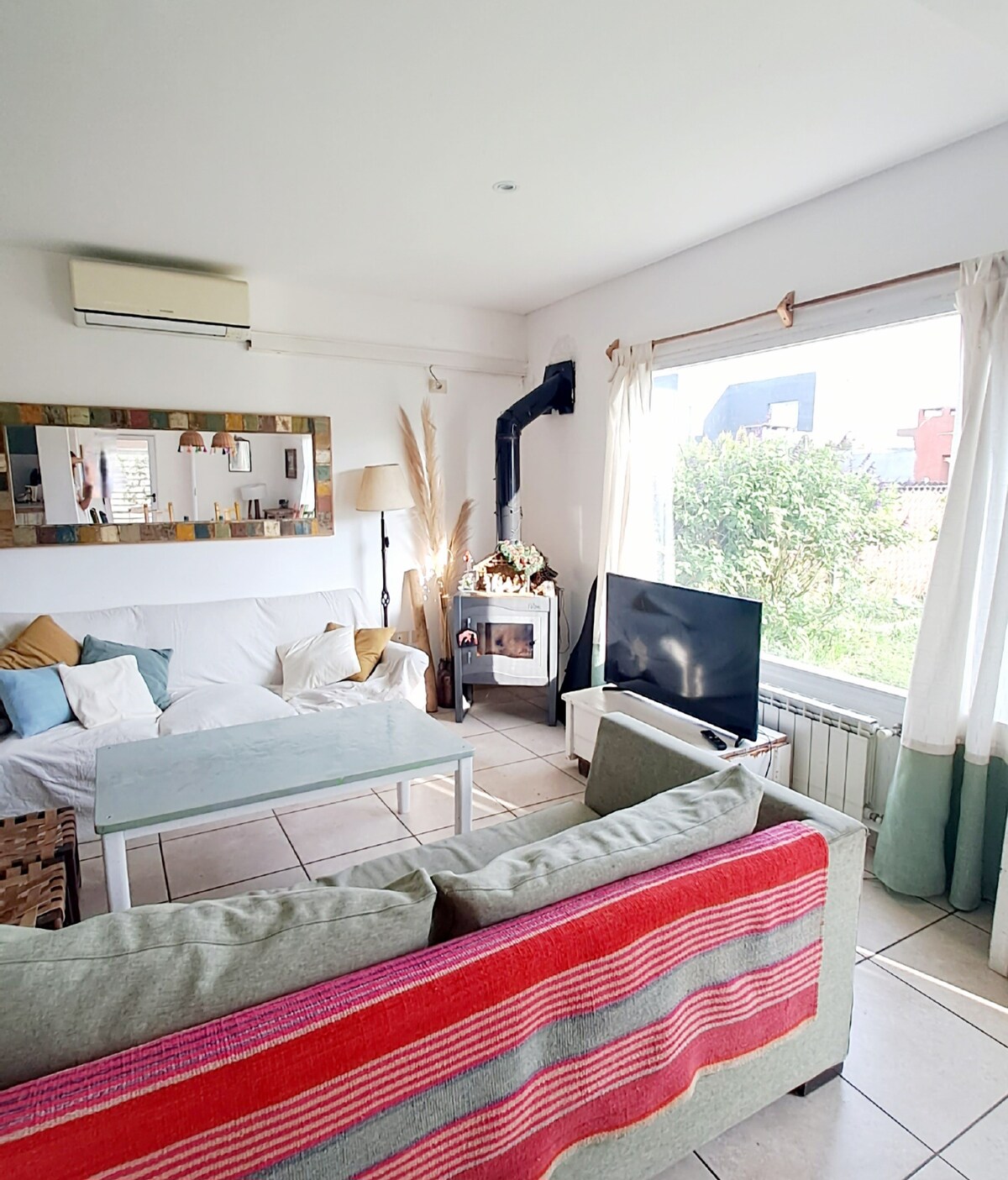 A bright living area features a comfortable white sofa adorned with decorative cushions. A glass coffee table is positioned in front, while a television stands beside a window showcasing a view of greenery. A cozy fireplace is also visible, complemented by natural light filtering in.