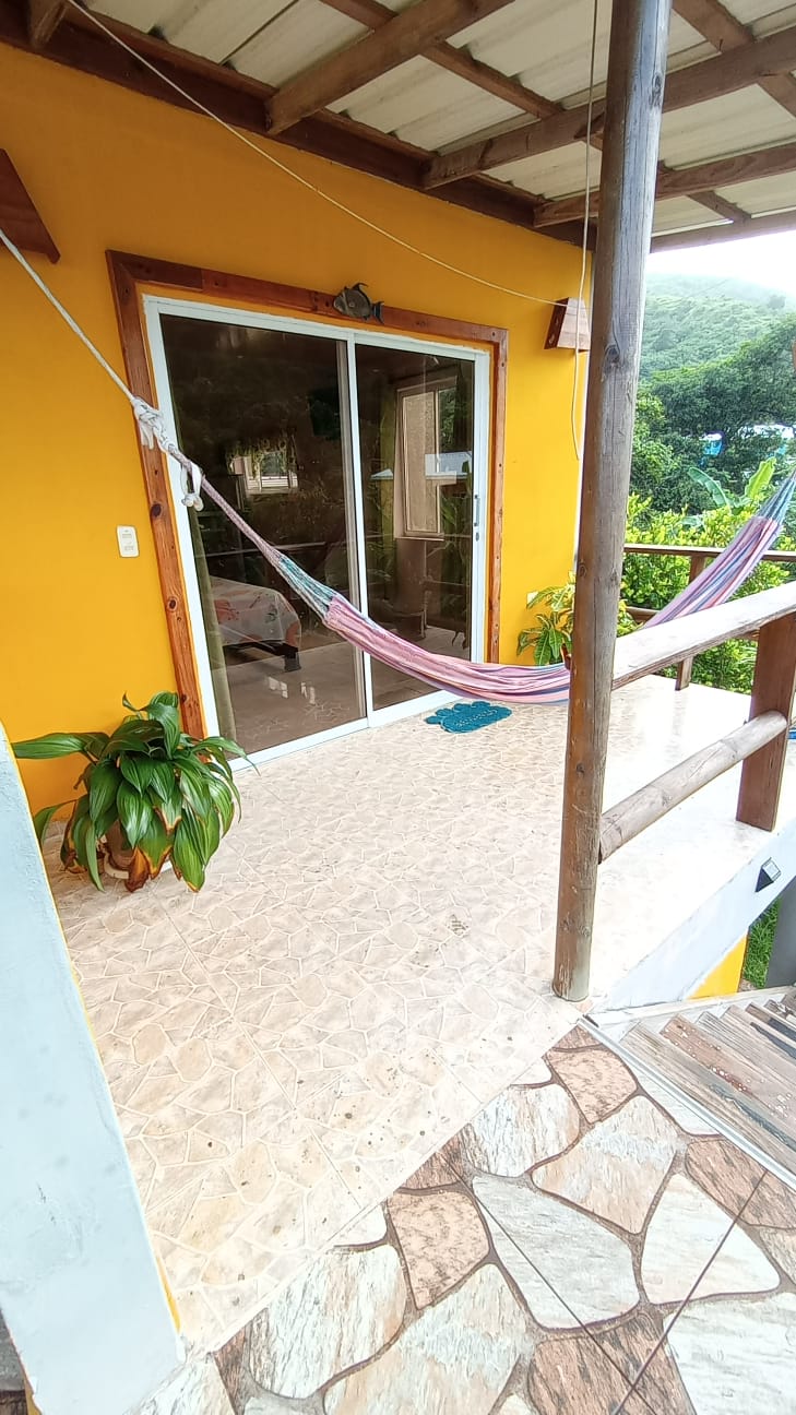 A covered balcony features a hammock strung between wooden posts. Potted plants are placed near the entrance, and floor tiles display a natural stone pattern. Large glass doors provide access to the interior, framed by warm yellow walls.