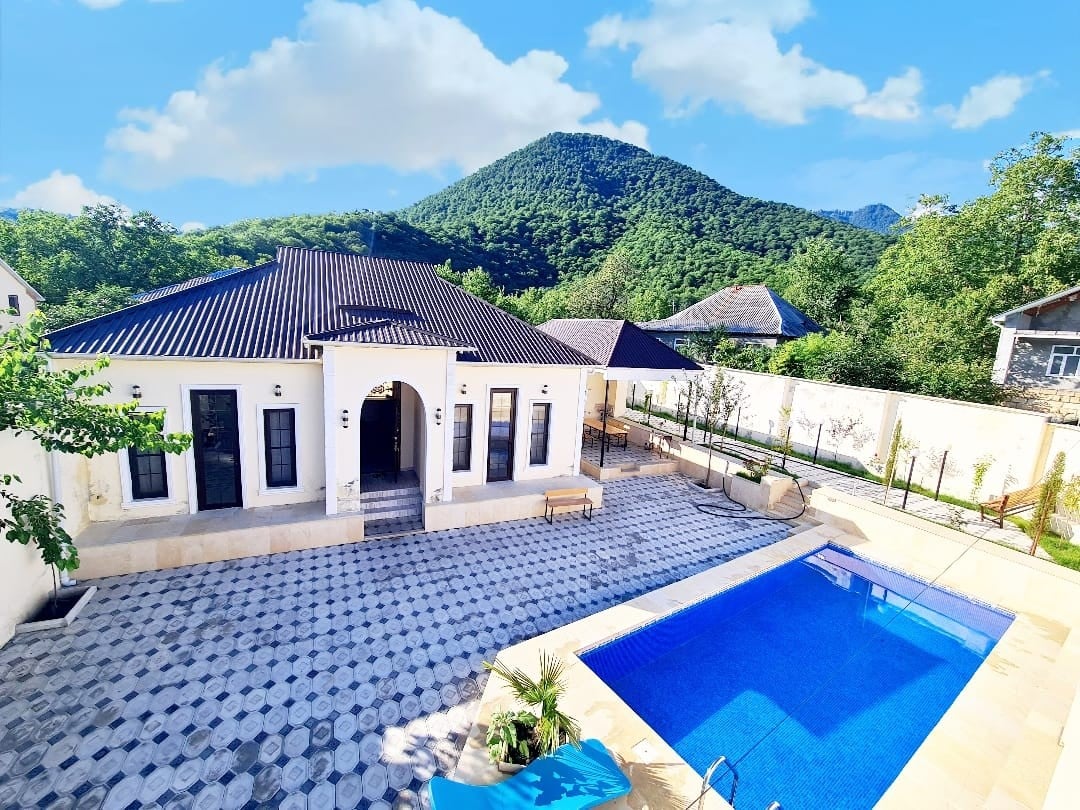 A one-story luxury villa is surrounded by lush greenery and mountains, featuring a spacious outdoor area with a blue swimming pool. The building's architecture is complemented by a patterned stone terrace, while large windows invite natural light into the interior.