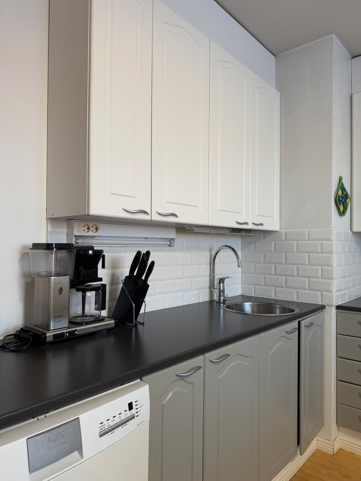 A modern kitchen features sleek cabinetry in white and grey, with upper cabinets providing ample storage. A black countertop hosts a coffee maker and utensils in a holder. A stainless-steel sink is positioned near the tiled backsplash, offering a clean and functional cooking space.