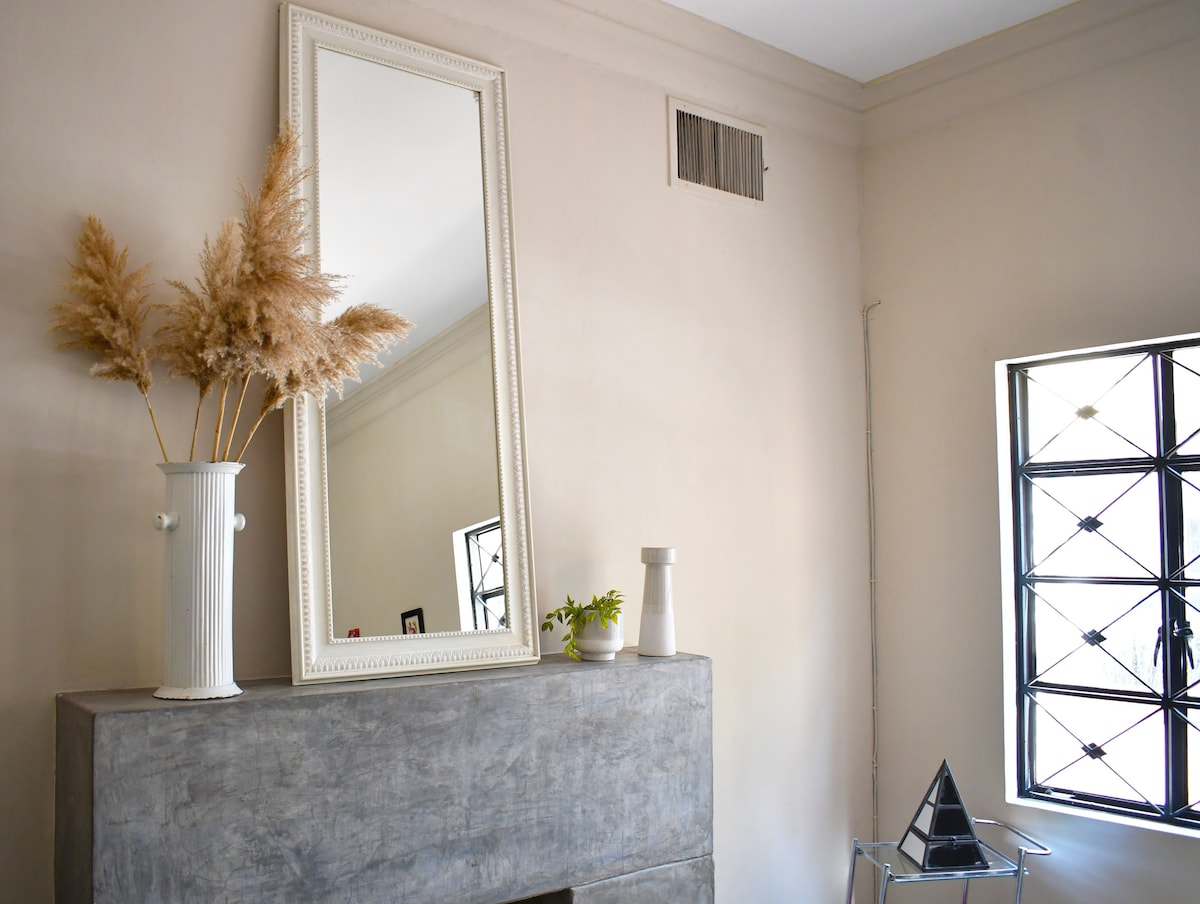 A neutral wall is complemented by a large mirror with a decorative frame, reflecting the light. A white vase holds dried pampas grass, while another minimalist vase sits nearby. A geometric sculpture adds a modern touch, and a window features a grid pattern of black bars.