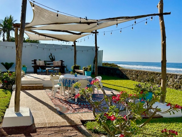 An outdoor seating area is framed by a wooden pergola, adorned with string lights. A colorful tablecloth covers a dining table surrounded by chairs, with a backdrop of manicured grass leading to the beach. The ocean is visible beyond a low stone wall, enhancing the coastal vibe.