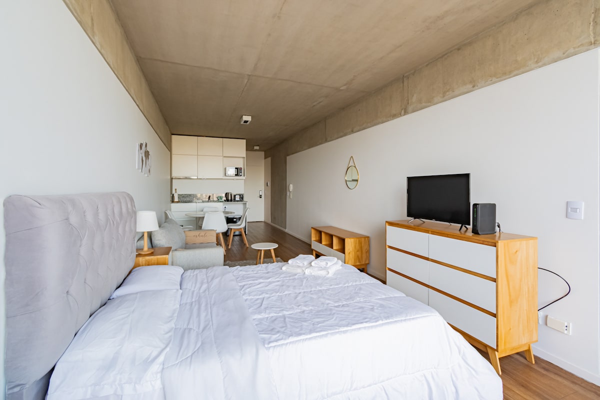 A modern studio apartment is presented, featuring a spacious bed with a light-colored headboard. In the background, a dining area with a table and two chairs is visible, along with a kitchenette. A TV sits atop a low wooden cabinet, providing a clean and minimalist aesthetic.
