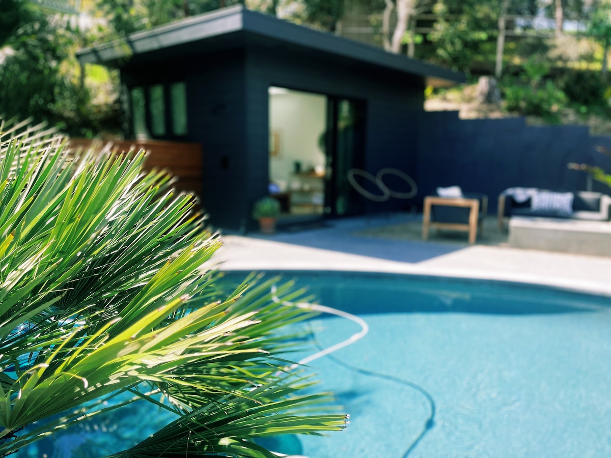 A serene pool area is framed by lush greenery, with the water reflecting light. The bathhouse features large glass doors, providing an inviting entry point. Comfortable outdoor seating is positioned nearby, creating a peaceful space for relaxation.