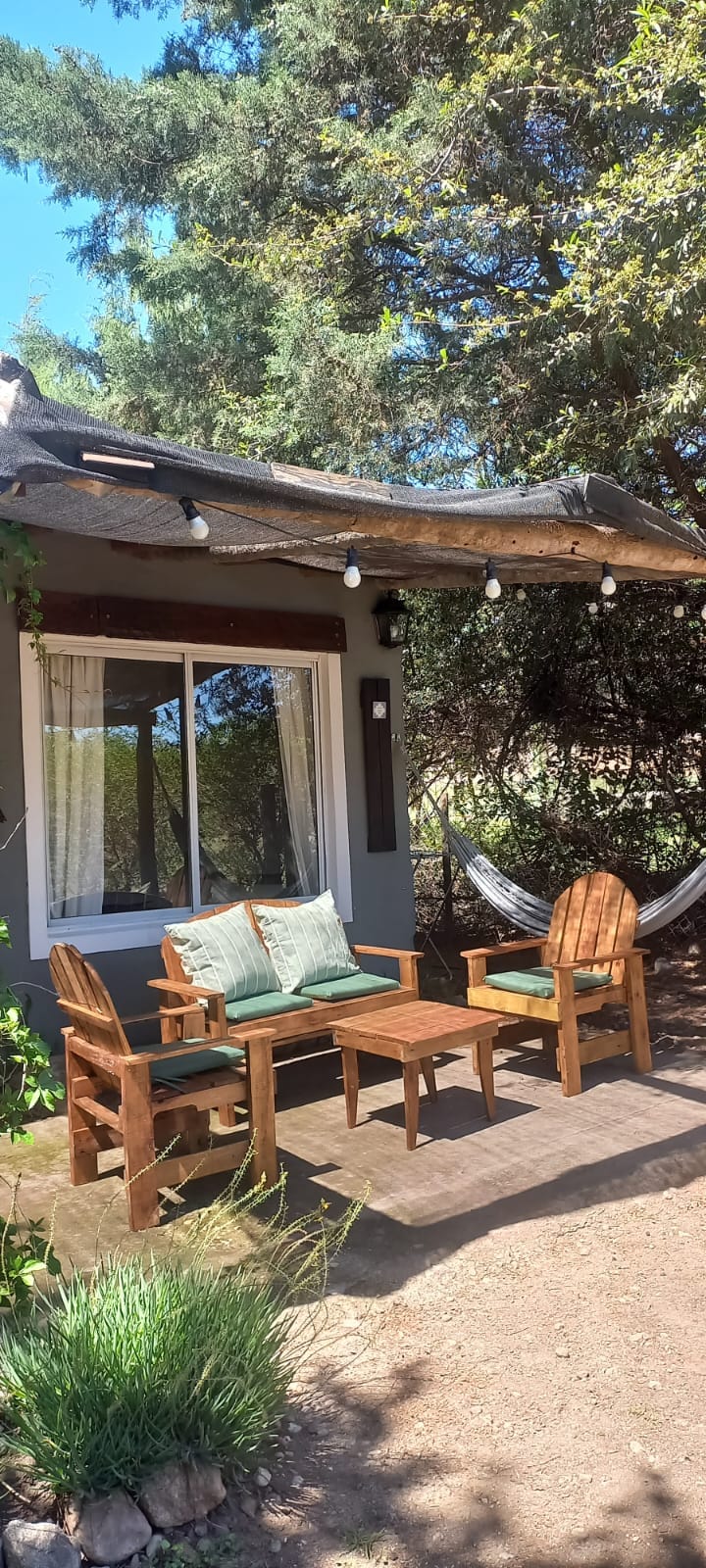An outdoor seating area features wooden chairs with green cushions and a small table, surrounded by lush greenery. A hammock is gently swaying between two trees, and string lights are draped overhead, enhancing the inviting atmosphere of the serene retreat.