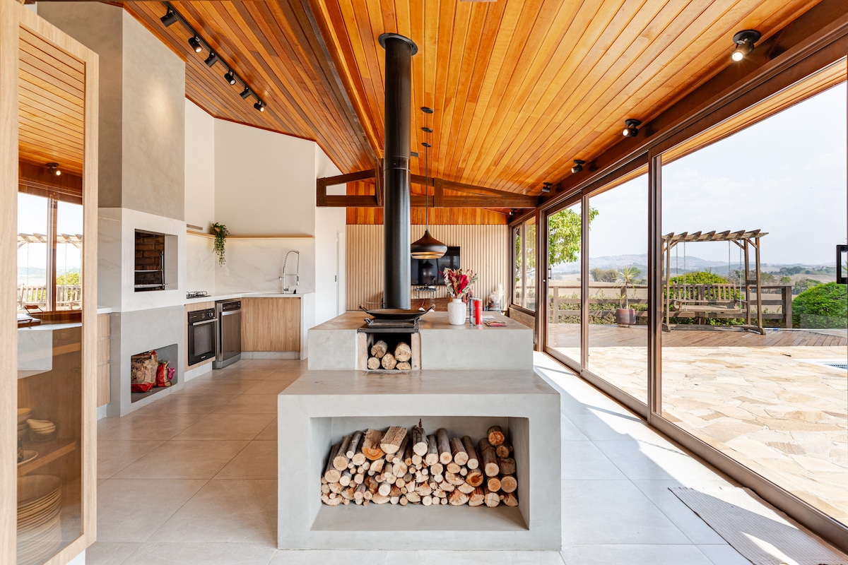 A modern kitchen space features a central cooking area with a wood storage compartment, complemented by large glass windows that offer expansive views of the outdoors. The wooden ceiling enhances the natural ambiance, while appliances are neatly integrated into the design.