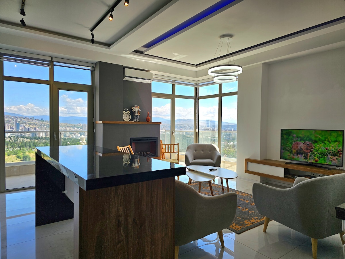 A modern living room is presented, featuring a dark countertop island that opens to a spacious area. Two stylish armchairs and a comfortable sofa frame a coffee table, while a large Smart TV and an electric fireplace add warmth. Expansive windows offer sweeping views of the surrounding landscape.