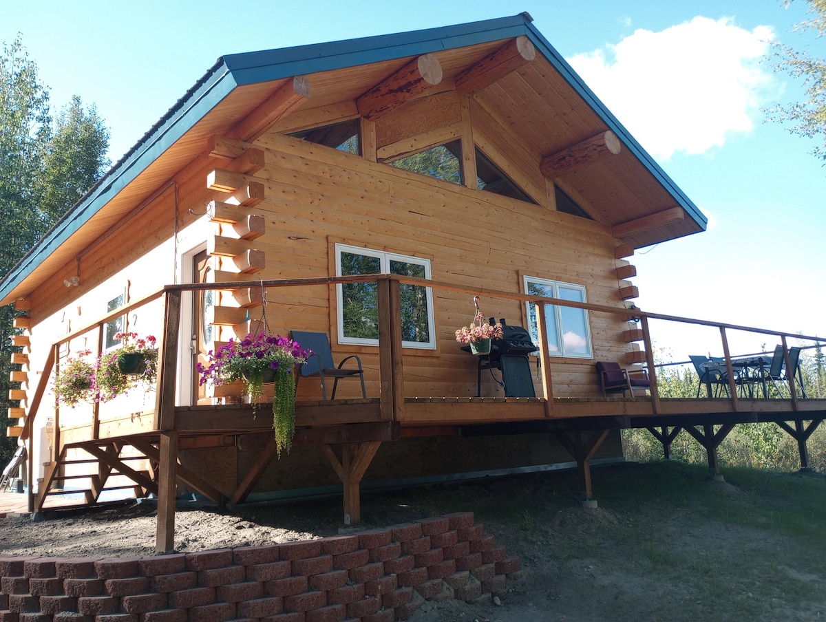 A newly-built log home is showcased from a low angle, featuring a spacious wooden deck adorned with colorful flowering plants. Large windows reflect natural light under a blue sky, while the distinct wooden architecture contributes to a welcoming exterior.