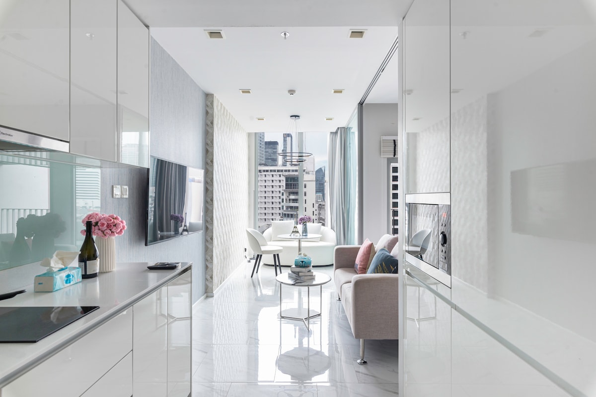 A modern living space is visible, featuring sleek, reflective surfaces and ample natural light. A cozy seating area with a light-colored sofa and a round coffee table complements a dining area with stylish chairs. Large windows offer views of the cityscape.