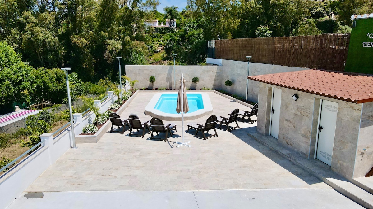 A private outdoor area features a swimming pool surrounded by a spacious patio. Several lounge chairs are arranged for relaxation, with umbrellas offering shade. The setting is enhanced by green foliage and a stone wall, creating a serene environment for outdoor enjoyment.