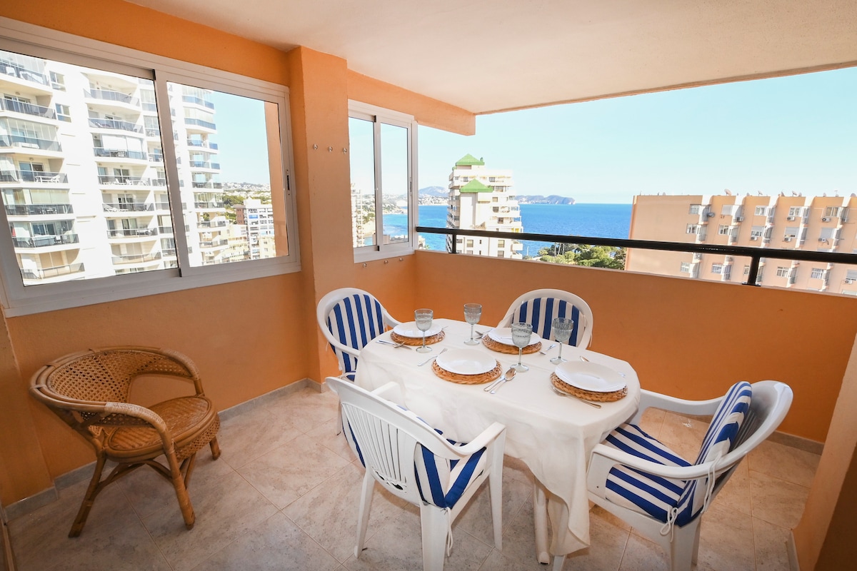 A large terrace is displayed with a dining table set for four, featuring white tableware and a tablecloth. Two chairs with striped cushions and a rattan chair enhance the outdoor space. Panoramic views of the sea and surrounding buildings are visible through large glass windows.