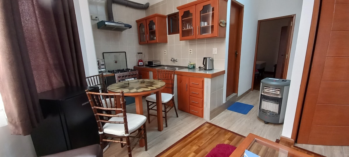 An open kitchen and dining area feature wooden cabinetry and a round dining table with four chairs. Kitchen appliances include a stove, kettle, and refrigerator. Natural light enters through a nearby window, illuminating the space. A small heater is positioned in the corner, enhancing comfort.