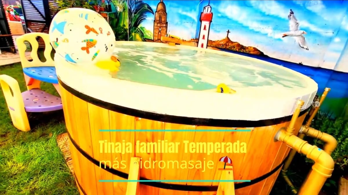 A family-sized, electronically heated hot tub is featured in a wooden barrel design. Colorful floats are visible on the surface of the water, surrounded by a grassy area. A vibrant mural depicting a lighthouse and seagulls enhances the relaxing atmosphere.