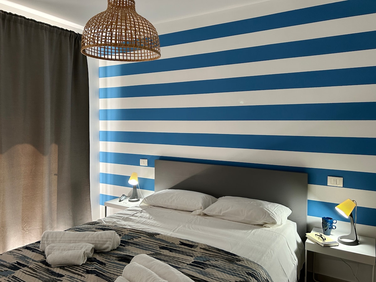 A bedroom is depicted with a striped blue and white accent wall. A spacious bed is positioned with two white pillows and fresh linens. Soft lighting is provided by lamps on each side, complemented by a textured woven light fixture above. Dark curtains frame a window.