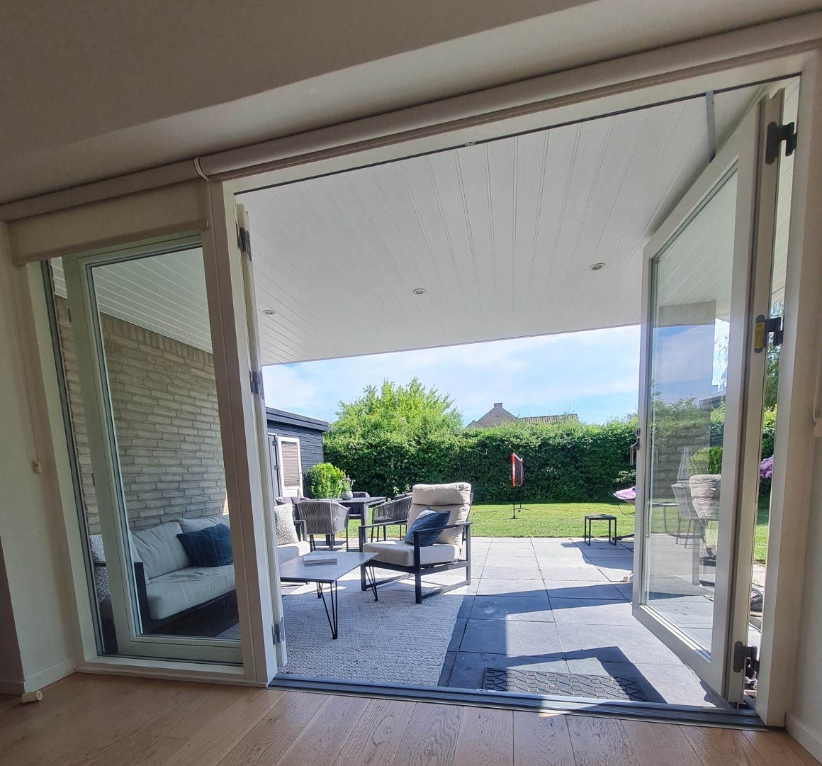 The image captures a seamless transition to an outdoor terrace featuring a seating area with cushions and a dining table. Large glass doors invite natural light, providing a view of the well-manicured garden and greenery beyond, enhancing the sense of openness.