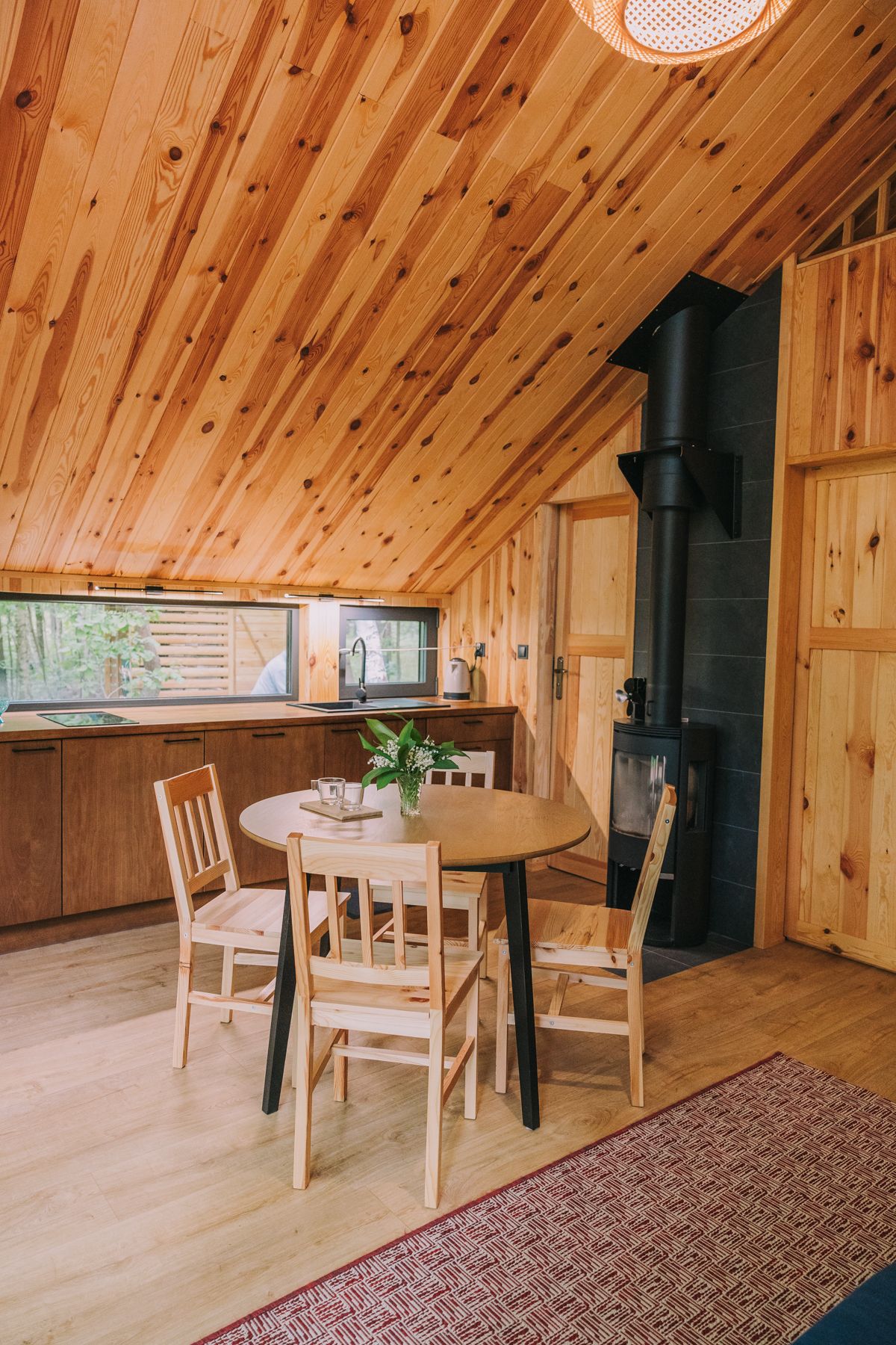 A bright dining area features a round wooden table surrounded by four light-colored chairs. Large windows allow natural light to fill the space, complemented by the warm wooden ceiling. A small plant sits on the table, adding a touch of greenery.