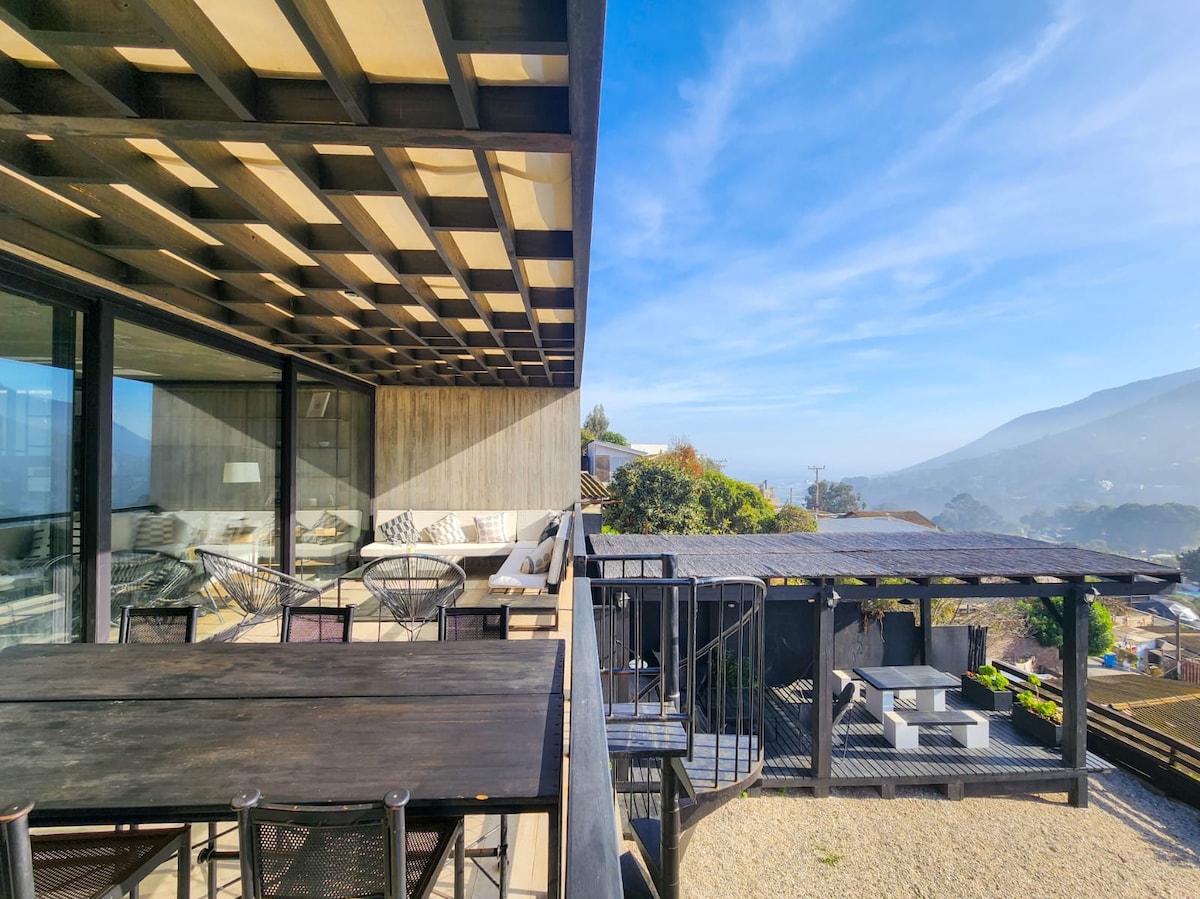 An outdoor terrace features a spacious wooden table surrounded by dark metal chairs. Lounge seating is positioned under a covered area, offering views of distant hills and a clear sky. Sunlight casts soft shadows, enhancing the outdoor experience.