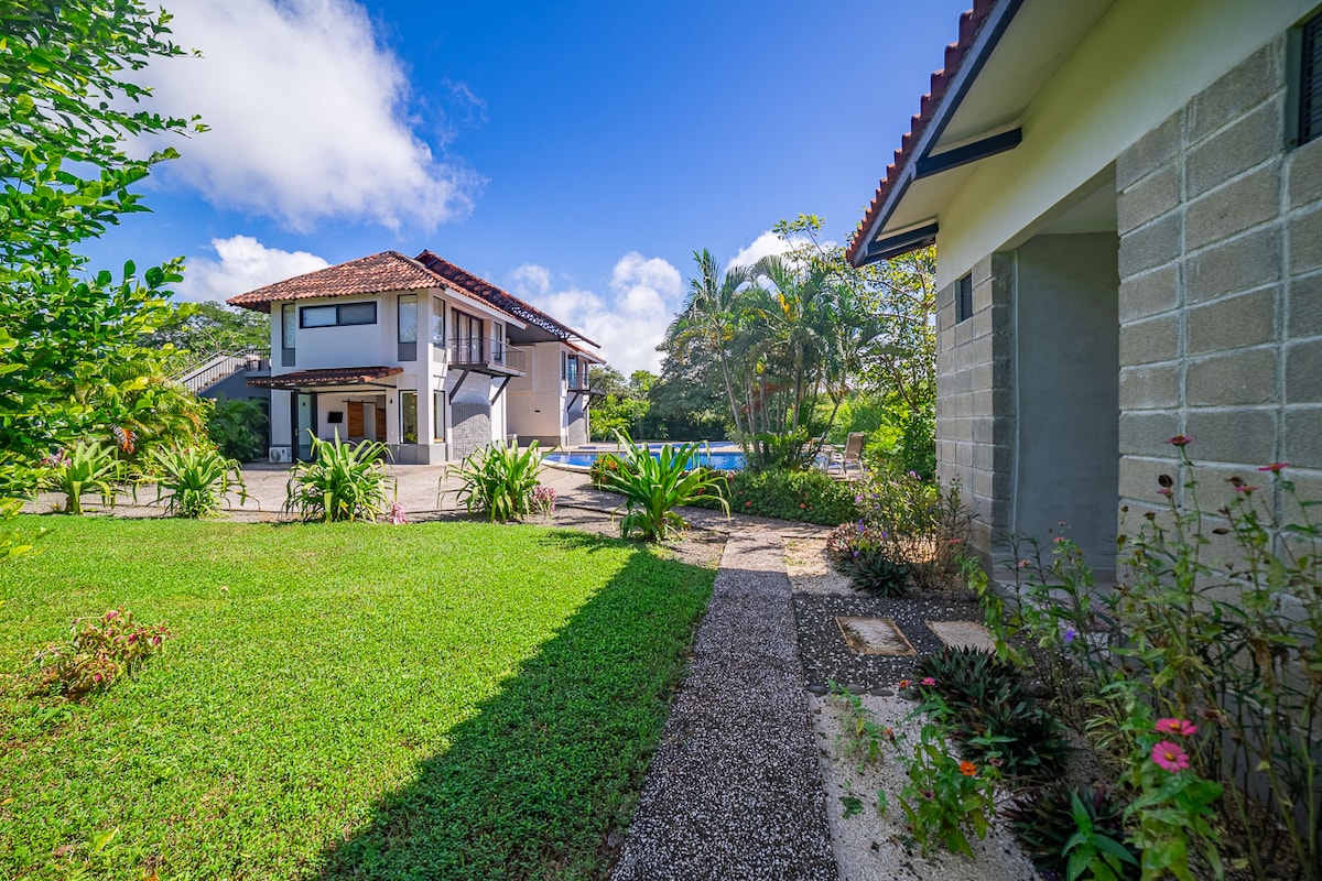The image captures a lush green pathway leading to a serene outdoor space. The well-maintained garden features vibrant plants and flowers, while a modern two-story building and a pool are visible in the background under a clear blue sky.