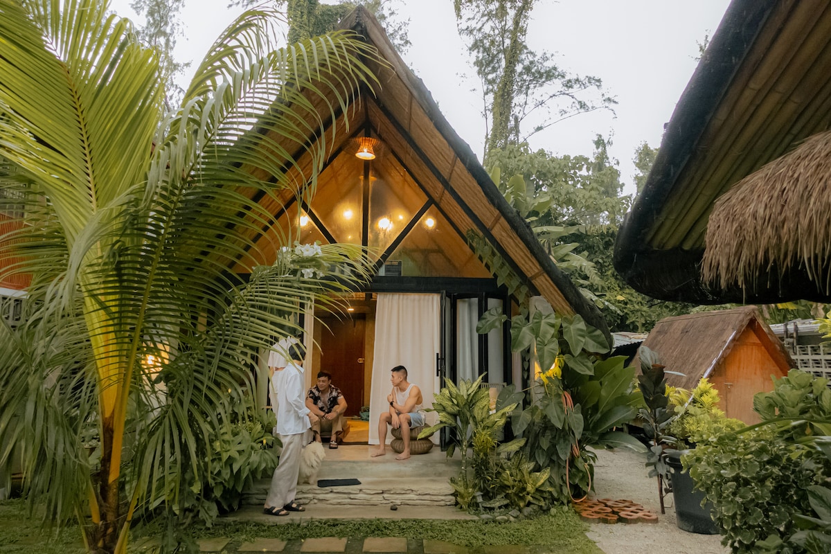 The entrance to the villa is framed by lush greenery, featuring a welcoming porch area with tropical plants. A cozy seating arrangement is visible, inviting relaxation. The unique A-frame roof design complements the tranquil atmosphere, while soft lighting enhances the overall inviting aesthetic.