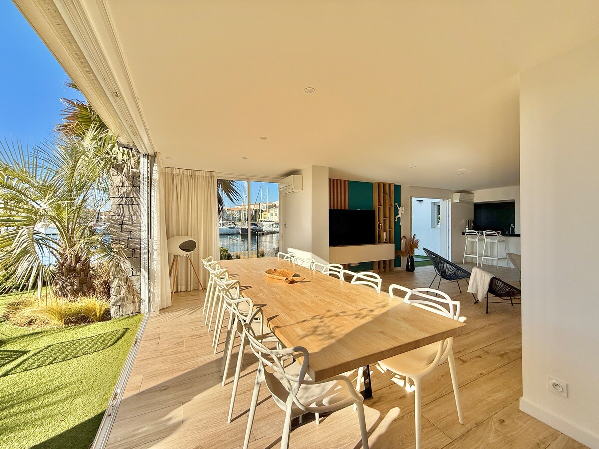 A bright and airy dining area is showcased with a long wooden table surrounded by white chairs. Large glass doors offer views of the marina, while palm trees add a touch of greenery. The space flows into a cozy lounge area visible in the background.