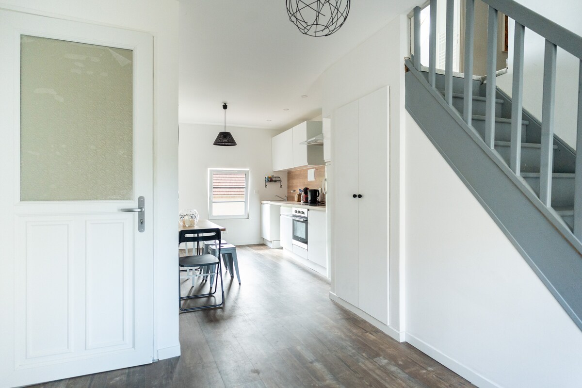 An open and airy entryway leads to a well-equipped kitchen and dining area. Soft natural light filters through a window, illuminating a wooden dining table surrounded by black chairs. A staircase is visible, featuring a light grey railing, enhancing the modern design.