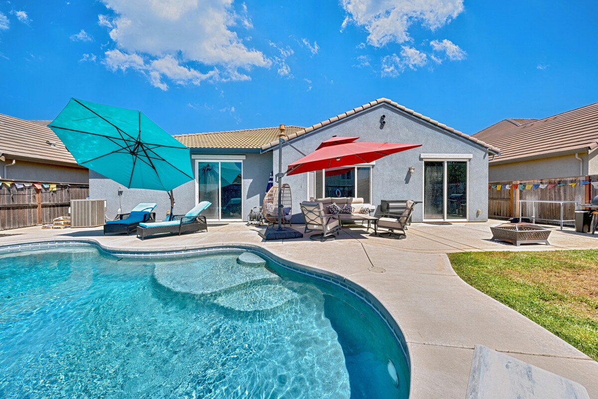 The backyard features a sparkling swimming pool surrounded by a sun-soaked patio. Colorful umbrellas provide shade over lounge chairs and a dining area. A well-kept lawn is visible, and the home's exterior showcases a welcoming design under a clear blue sky.