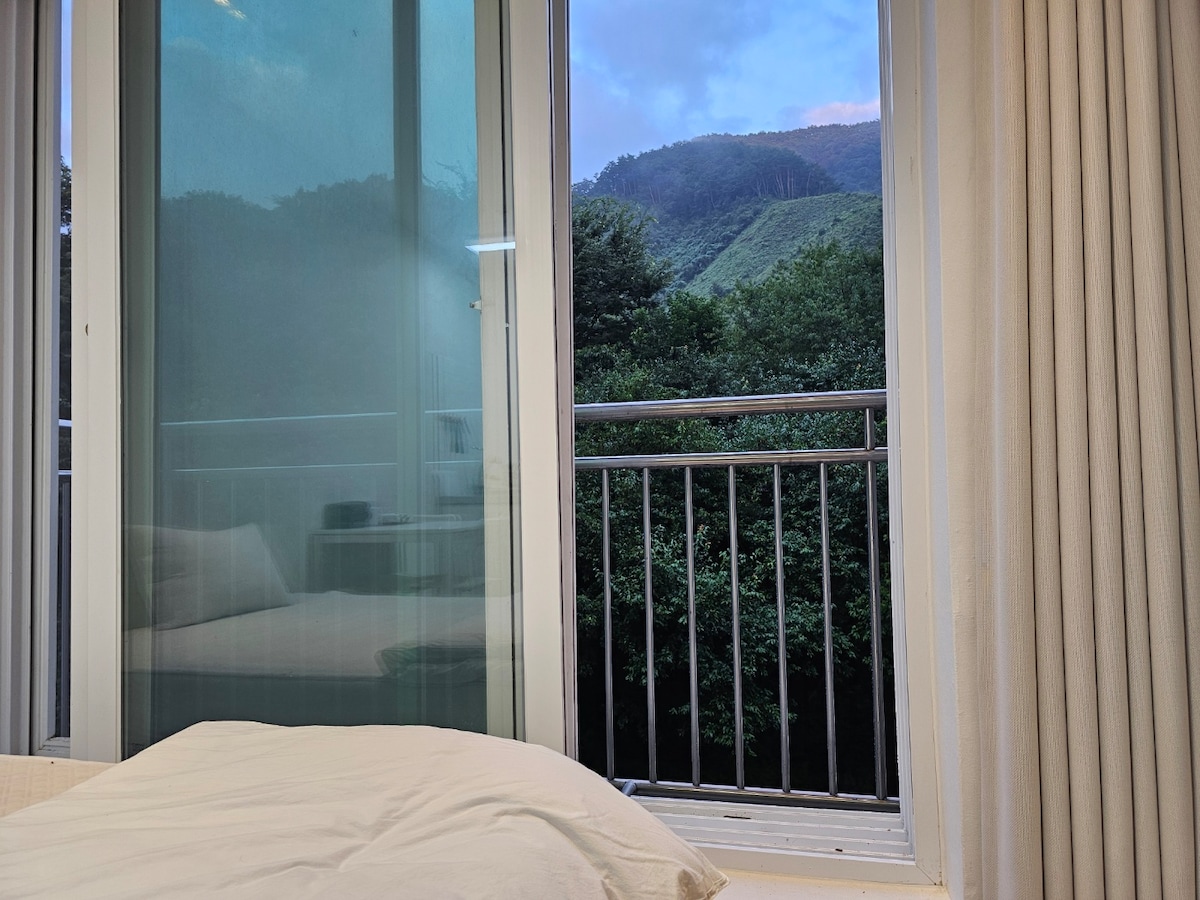 A view from the bedroom showcases a sliding glass door that opens to a balcony. Lush greenery is visible outside, complementing the serene mountain landscape beyond. Soft curtains frame the view, while a spacious bed rests in the indoor setting.