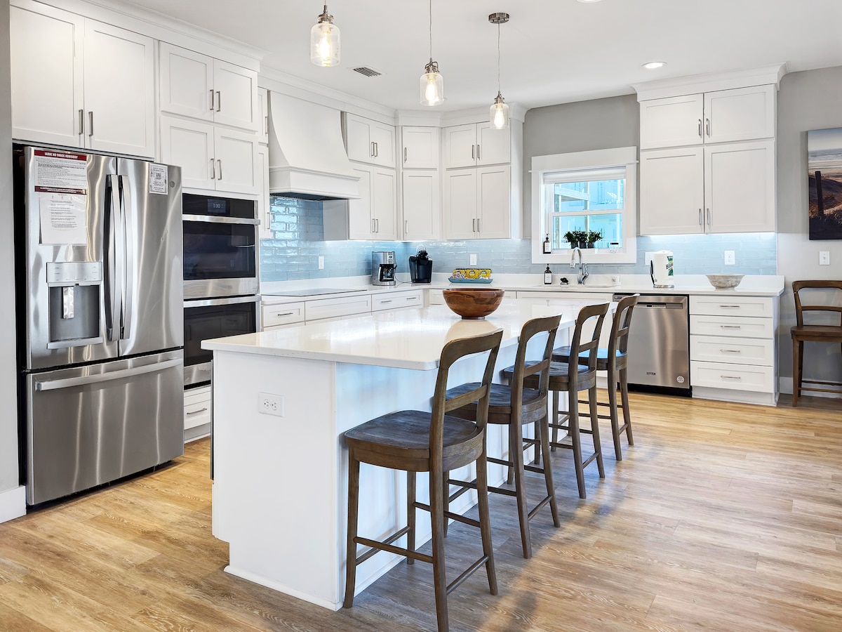 A modern kitchen features white cabinetry and stainless steel appliances, including a spacious refrigerator and double ovens. A large island with seating for six is illuminated by pendant lighting, providing a bright, inviting atmosphere. Natural light enters through a window, enhancing the clean design.