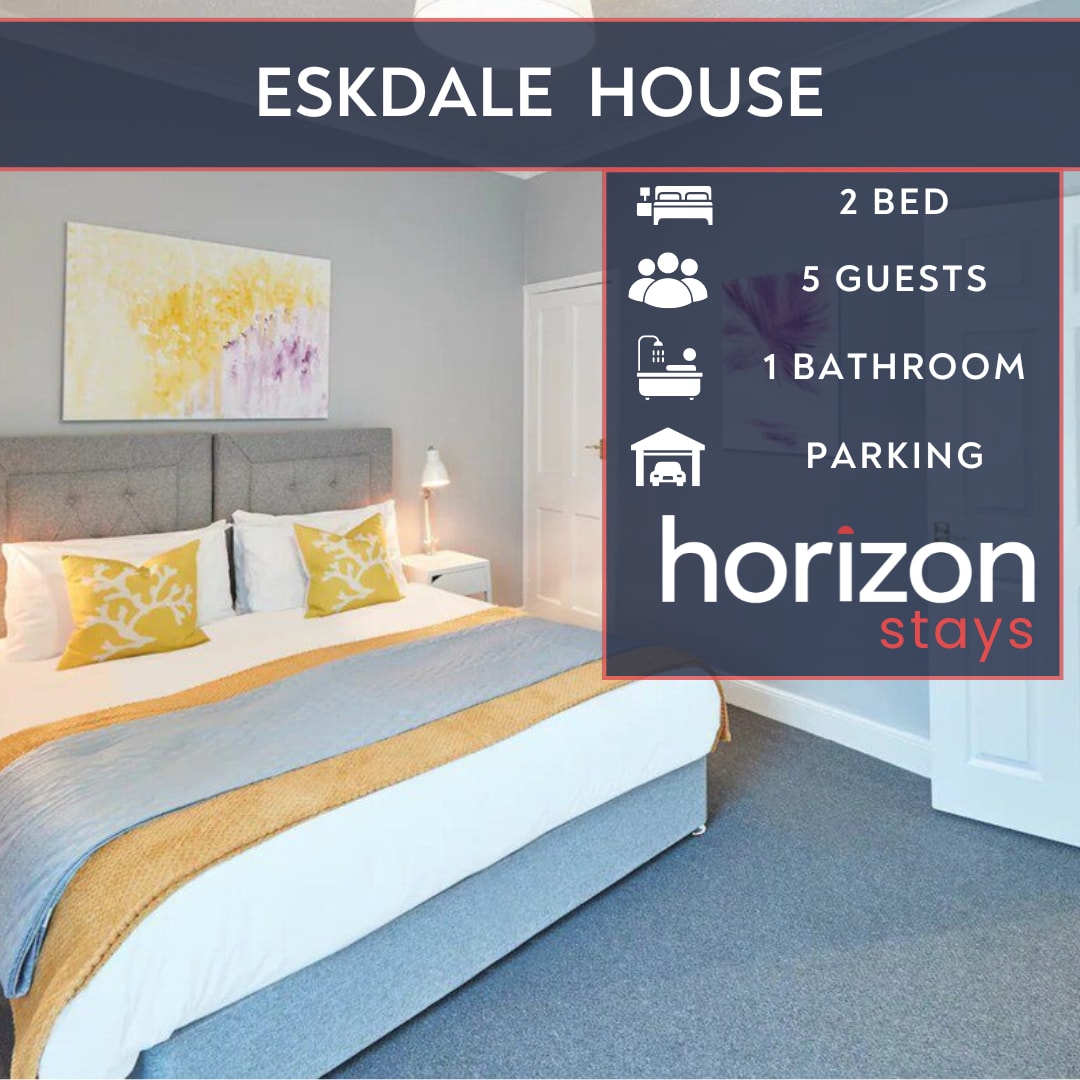 The bedroom features a spacious bed with a light grey headboard and bright yellow accent pillows. A soft gray carpet covers the floor, while a colorful piece of wall art adds a touch of design. Natural light enters through the window, creating a calm ambiance.