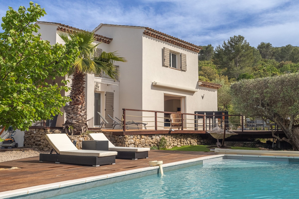 A two-story house is situated on a hillside, surrounded by lush greenery. A spacious deck extends from the house, featuring lounge chairs positioned near a pool. A palm tree adds to the tropical feel, enhancing the natural landscape.