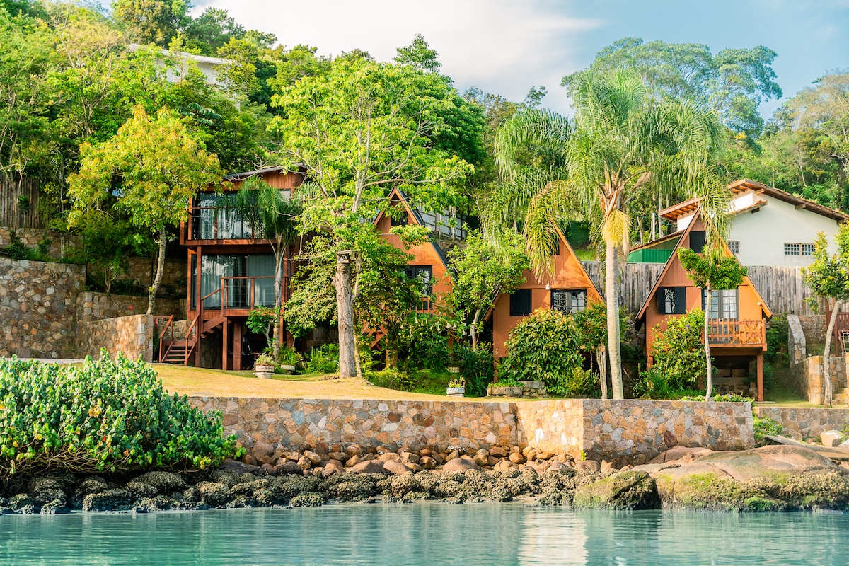 A charming collection of rustic chalets is set against a lush green backdrop, harmonizing with the natural landscape. Palm trees and shrubs enhance the inviting exterior, while the clear water reflects the colorful elements of the chalets. The tranquility of the environment is evident.