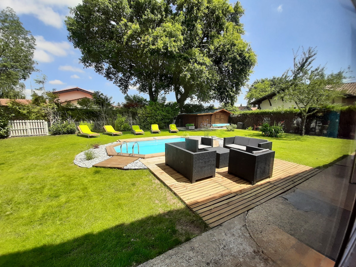 A spacious backyard is presented, featuring a private pool surrounded by a wooden deck. Comfortable seating arrangements are located adjacent to the pool. Sun loungers are positioned for relaxation, while greenery and trees enhance the outdoor setting. A peaceful atmosphere is evident in this inviting space.