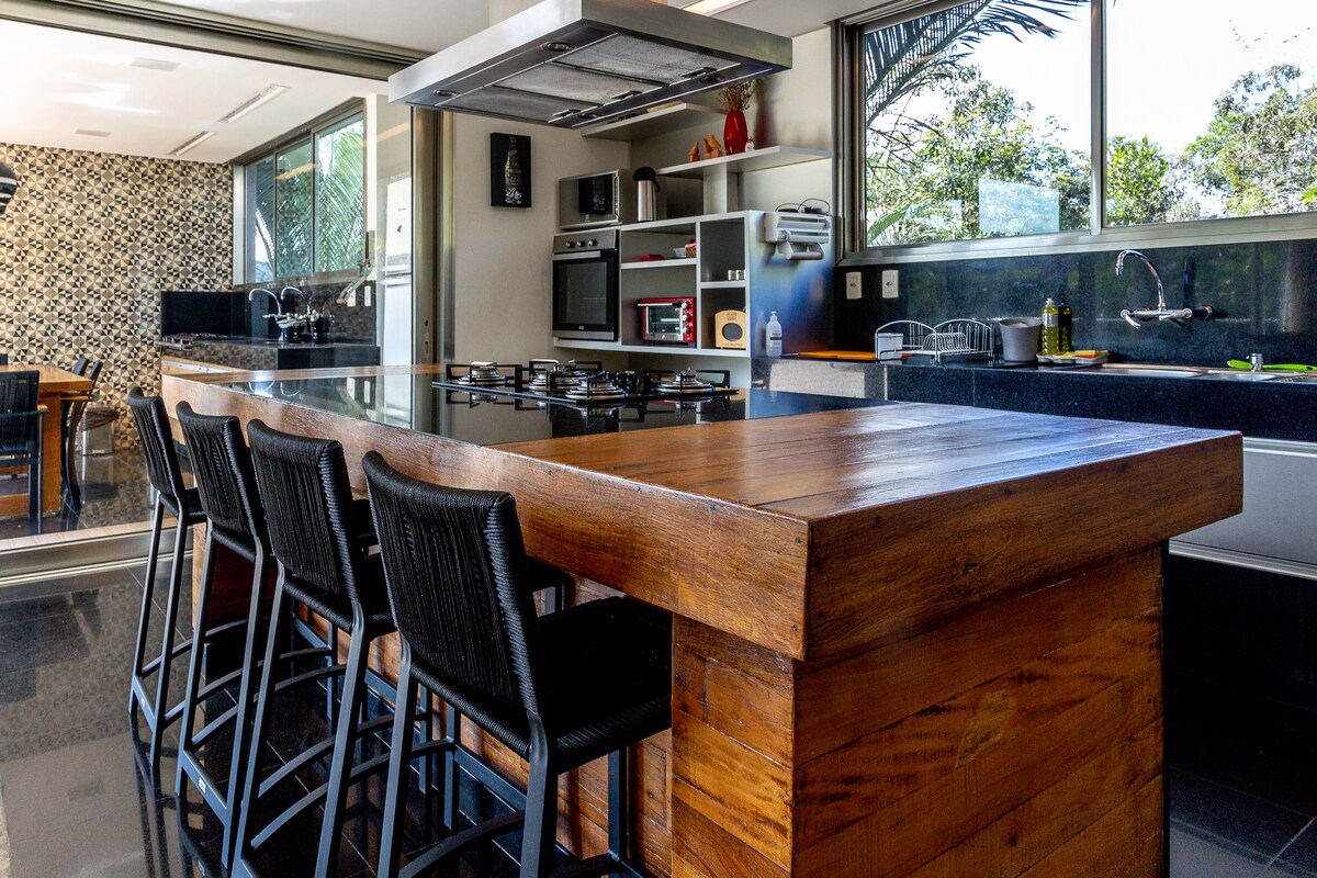 A modern kitchen is shown with a large wooden island featuring bar stools, equipped with a built-in stovetop and range hood. Storage cabinets are visible above and below counter surfaces, with appliances neatly arranged. Natural light enters, highlighting the dark countertops and surrounding greenery.